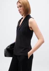 Μαύρο αμάνικο τοπ με V-λαιμόκοψη, κατασκευασμένο από απαλό ύφασμα. Διαθέτει άνετη εφαρμογή και διακριτικό draping στον λαιμό. Φορώντας το με μαύρο παντελόνι.