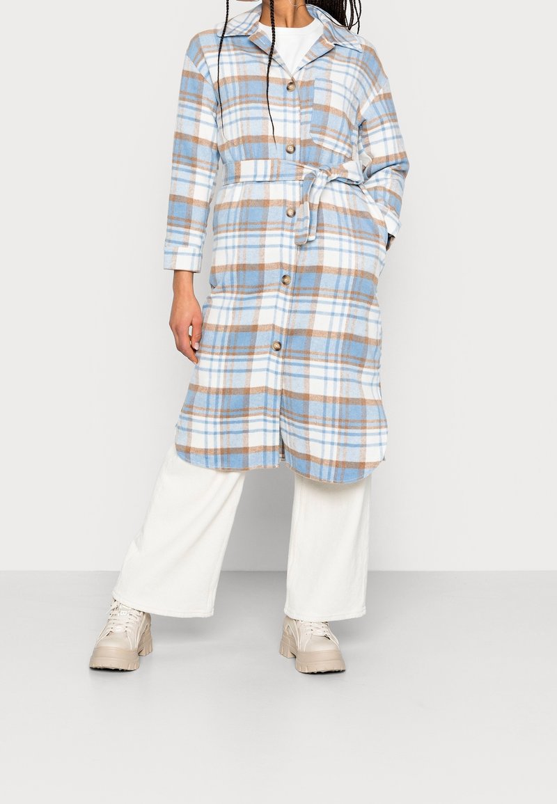 Long manteau à carreaux bleu clair, blanc et beige avec boutons et ceinture à la taille, porté avec un pantalon blanc à jambes larges et des chaussures beiges épaisses.