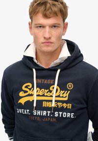 Marineblaues Hoodie mit weißen Kordeln und übergroßem Logo in Gelb und Braun. Der Stoff hat eine weiche Textur; schichtartiger Kragen in Weiß.