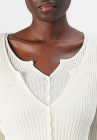 Lys farget ribbestrikket cardigan med splitte halslinning, teksturert stoff og dekorative knapper nedover midten.