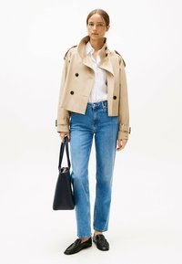 Abrigo de trench corto beige con botones de doble botonadura, camisa blanca, jeans rectos azules y bailarinas negras. Sosteniendo un bolso oscuro.