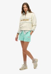 Biała bluza z pół zamek błyskawiczny z logo "SUPERDRY", zestawiona z miętowozielonymi szortami z lekkim przetarciem i logiem na kieszeni.