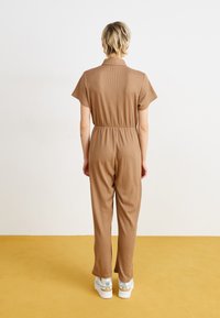 Combinaison marron côtelée avec des manches courtes, taille cintrée et jambes droites. Portée avec des baskets blanches et bleues, sur un sol jaune.
