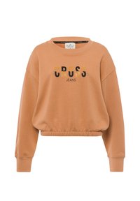 Jasnobrązowy krótki sweter, z okrągłym dekoltem, luźnym krojem oraz czarnym napisem z złotymi akcentami, który brzmi "CROSS JEANS."