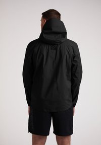 Mand iført en sort hættejakke og sorte Under Armour shorts, vendt væk mod en neutral baggrund.