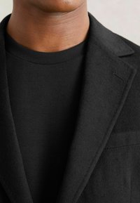 Sort skræddersyet jakke med en blød tekstur og klapkrave, lagdelt over en sort rundhalset t-shirt, der fremhæver et stilfuldt monokrom look.