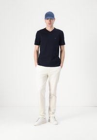 Tamnoplavi pamučni T-shirt s V-izrezom, bijele hlače i bijele tenisice, uparen s plavom kapom. Model stoji uspravno, ruke u džepovima.