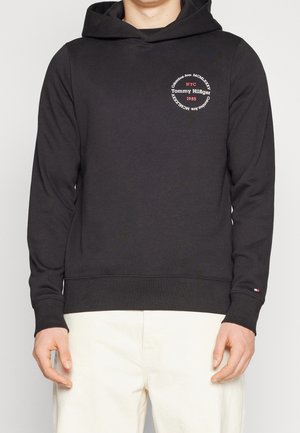 Mężczyzna w czarnej bluzie z kapturem Tommy Hilfiger z okrągłym białym i czerwonym logo na piersi oraz małym logo flagi na mankiecie lewego rękawa.