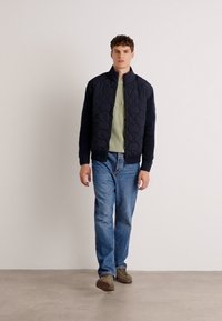 Marineblå, quiltet jakke med oppreist krage, båret over en lys grønn stripete skjorte, kombinert med blå jeans og brune sko. Glatt tekstur.