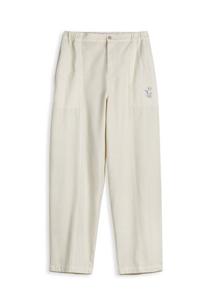 Mujer con blusa sin mangas amarilla de cuadros, pantalones crema de talle alto y zapatos planos plateados metálicos, de pie contra un fondo liso.