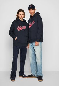 Marineblå hettegensere med dristig rød logo, kombinert med marineblå bukser med hvite striper og lys blå denim, med matchende mørke joggesko og caps.