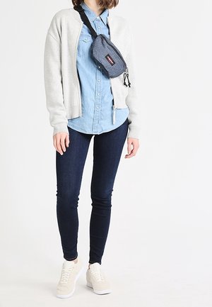 Persoon draagt een lichtgrijs jack met rits, een blauw overhemd met knopen, donker strakke jeans, beige sneakers en een grijze schoudertas.