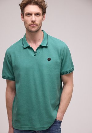 Mężczyzna o krótkich, kręconych włosach, ubrany w zieloną koszulkę polo z czarnymi wykończeniami i małym czarnym okrągłym logo na piersi, ręce w kieszeniach jeansów.