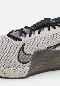 Nike sportiniai bateliai turi šviesiai pilką tinklelio viršutinę dalį, juodus raiščius, struktūruotą tamsiai pilką pado dalį su dėmėtu akcentu ir ryškiu logotipu.