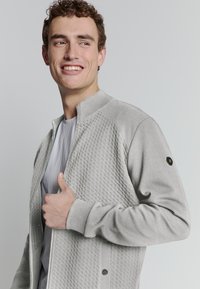 Jonge man die glimlacht, gekleed in een lichtgrijze textuur-ritsjack over een grijs shirt, poseert met één hand die de kraag van het jack vasthoudt.