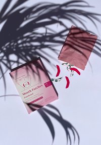 Emballage rose des "Patches pour la bouche à l'abricot" avec un design minimaliste, mettant en avant un dessin linéaire d'un visage. Des patches lâches en rose et avec des accents verts à motifs à proximité.