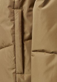 Brun vadderad jacka med detalj som visar en slät, texturerad yta, en sidoficka och synliga sömmar vid sömmarna.