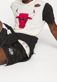 Krátký rukáv, krémové a černé bavlněné tričko s grafikou červeného býka a kruhovým logem NBA Finals. Nosené s černými síťovanými šortkami.