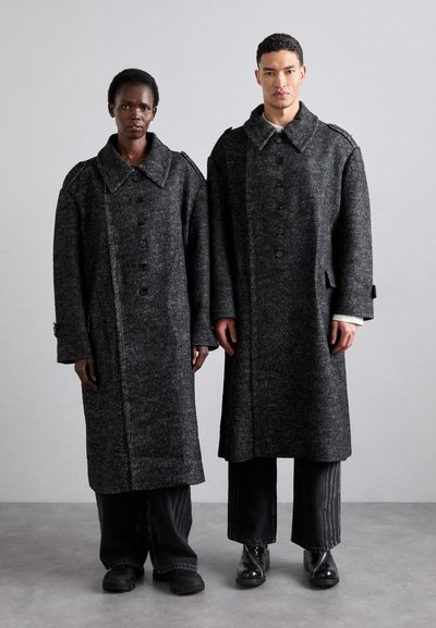 Abrigos largos, negros y texturizados con grandes cuellos y cierres de botones, llevados sobre pantalones oscuros de pierna ancha. Zapatos lisos y brillantes completan el look.