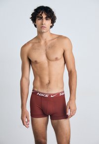 Brune Nike-bokser med hvit elastisk linning. Stoffet virker glatt med en tettsittende design. Modellen står skjorteløs mot en lys bakgrunn.