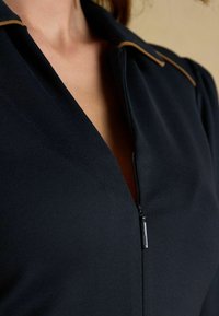 Tumesinise kleit V-kaelusega, millel on lukuga sulgemine ja pruun aktsent krae ääres. Sile kangatekstuur, sobiv disain.