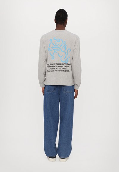 Gray long-sleeve shirt with blue graphic and text on the back. Paired with loose-fitting blue jeans and white sneakers.