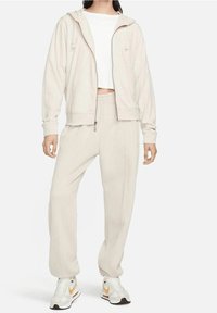 Beige zip-hoodie med dragsko, matchad med beige sweatpants. Båda är tillverkade av mjukt material, med en avslappnad passform och subtil textur.