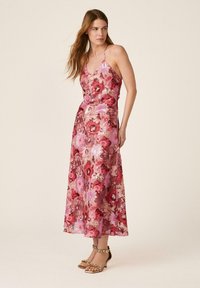 Femme debout dans une robe midi sans manches à fleurs roses et rouges avec fines bretelles, portant des sandales beige à talons hauts avec brides à la cheville.