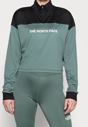 Femme portant un haut à manches longues The North Face vert et noir ainsi qu'un legging assorti avec le logo visible sur la poitrine et la cuisse.
