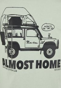 Bílé bavlněné tričko s černou grafikou vozidla s přístřeškem na střeše. Text zní „ALMOST HOME“ a „ANOTHER COTTON LAB“ s dekorativními detaily.