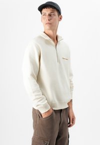 En ung mand med kort mørkt hår står og kigger eftertænksomt til sin højre, iført en cremegul zip-up sweatshirt og brune cargo-bukser.