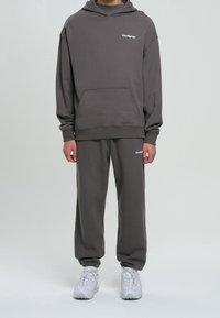 Sudadera gris con capucha y bolsillo frontal, combinada con pantalones de chándal a juego que tienen puños elásticos. Ambos presentan un pequeño logo blanco como acento.