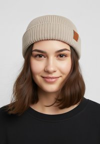 Beige ribbed knit beanie with a folded cuff and a small brown leather tag. Smooth texture; fitted design suitable for cold weather.