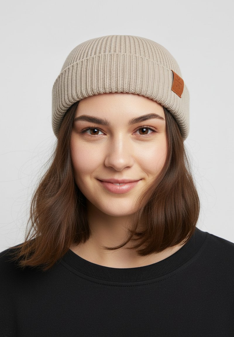 Beige ribbed knit beanie with a folded cuff and a small brown leather tag. Smooth texture; fitted design suitable for cold weather.
