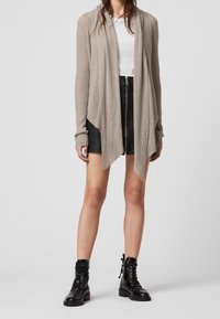 Cardigan de canalé beige claro con abertura frontal, manga larga y dobladillo asimétrico, combinado con una falda negra ajustada y botas negras con cordones.
