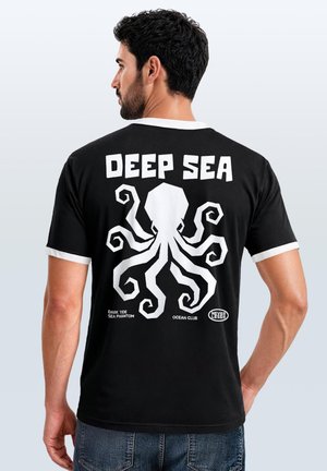 Mann im Seitenprofil trägt ein schwarzes T-Shirt mit weißem Krake-Grafik und Text "Deep Sea," "Dark Tide Sea Phantom," "Ocean Club," und "Nevertheless."