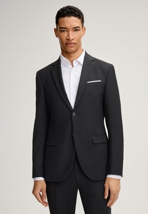 Uomo che indossa una giacca da abito nera aderente sopra una camicia bianca, in piedi contro uno sfondo chiaro e semplice, rivolto verso l'obiettivo.