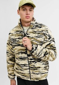 Chaqueta de forro polar en color marrón claro y negro con un patrón de rayas de tigre, cremallera completa, cuello alto y superficie texturizada. Diseño casual con bolsillos frontales.