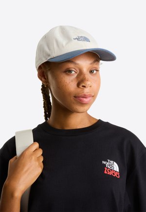 Světle šedá čepice s modrým kšiltem a vyšitým logem vpředu. Nošena s černým tričkem, které ukazuje hladkou texturu látky.