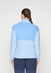 Lyseblå fleece-pullover med høy krage. Har et gult vannrett belte over ryggen. Myk tekstur, lange ermer og avslappet passform.