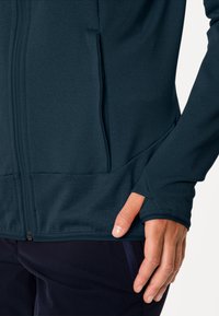 Giacca con zip di colore blu navy, con una texture liscia, cuciture piatte e fori per il pollice sui polsini. Presenta tasche laterali e un design aderente.