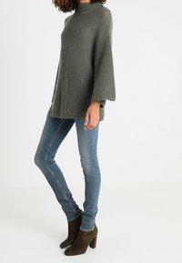Přehnaně velký, žebrovaný svetr v tmavě zelené barvě, s vysokým límcem a širokými rukávy. Nosený s modrými skinny džíny a hnědými kotníkovými botami na podpatku.