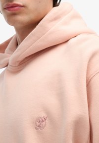 Sudadera con capucha de color rosa claro, hecha de tejido suave; presenta un pequeño logotipo bordado en un rosa más oscuro en la zona del pecho; ajuste relajado.