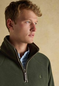 Giovane con capelli castano chiaro che indossa una giacca in pile verde scuro sopra una camicia a quadri blu, con lo sguardo rivolto a destra su uno sfondo semplice.