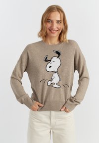 Pull en tricot beige avec un design de dessin animé Snoopy en noir et blanc sur le devant. Il a un col rond et des manches longues.
