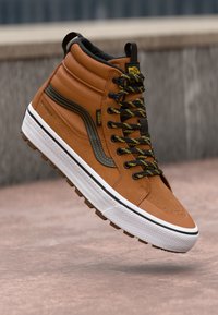 Korkeat ruskeat Vans-kengät, jotka on valmistettu vedenpitävästä materiaalista. Kenissä on mustat ja keltaiset nauhat, valkoinen pohja ja kuminen pitoalusta.