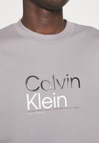 Felpa grigia con scollo rotondo. La parte anteriore mostra "Calvin Klein" in lettere nere e bianche in grassetto con una texture liscia.