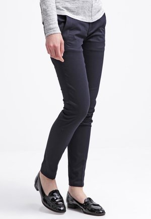 Person wearing fitted dark navy pants, gray long-sleeve top, and shiny black loafers standing with legs crossed.