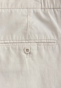 Tissu en coton beige avec une texture lisse, comportant un seul bouton blanc et une poche plate. Couture soignée le long des bords.