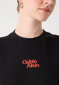 Svart bomulls t-skjorte med rød brodert "Calvin Klein"-logo på brystet, med rund hals og korte ermer.